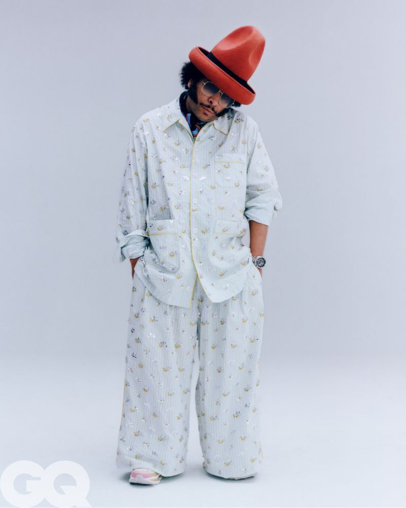 Boots Riley wearing a patterned light-colored shirt, matching wide-leg trousers, and a tall red hat against a white studio background.