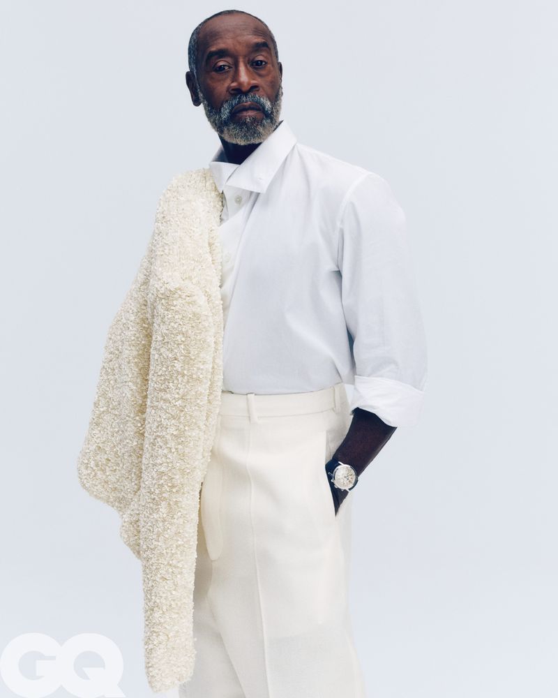 Don Cheadle wearing a white shirt and trousers, holding a textured jacket over his shoulder against a seamless white background.