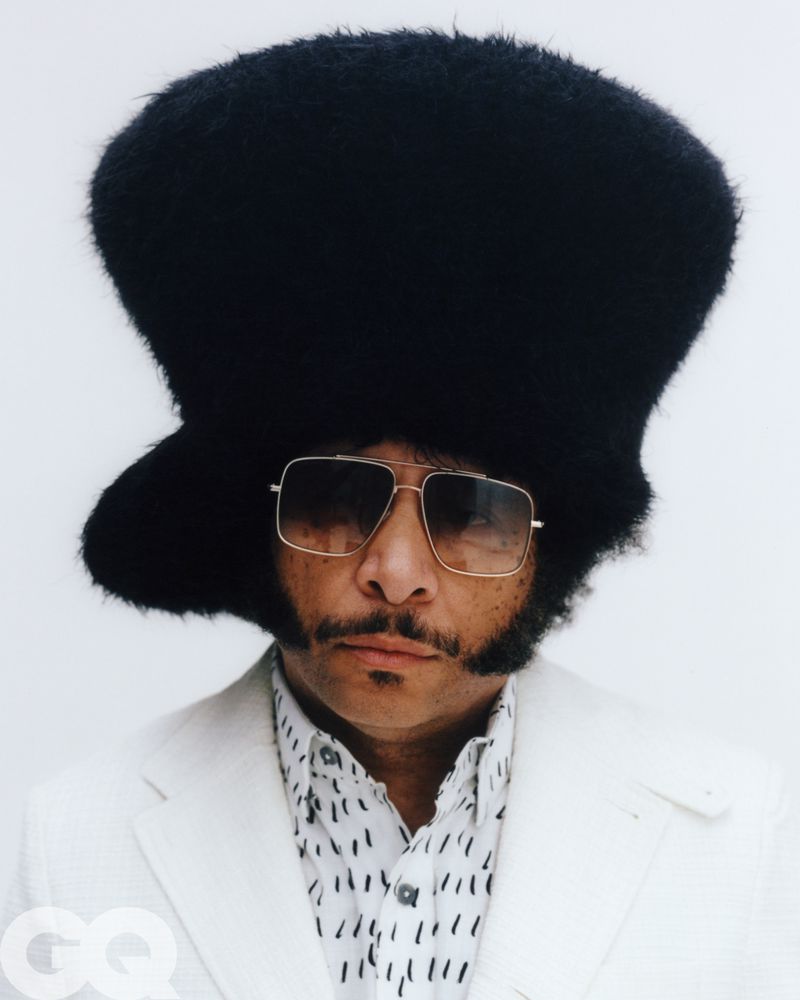Boots Riley wearing a large black furry hat, tinted sunglasses, and white suit jacket against a seamless white background.