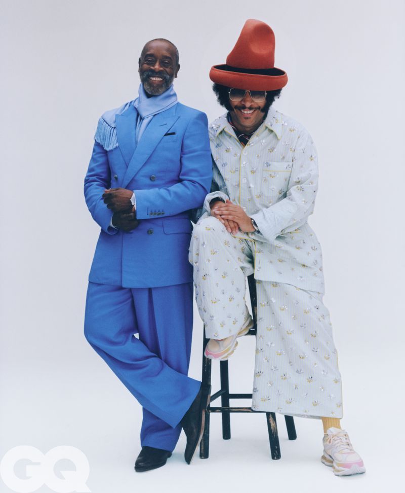 Don Cheadle in a blue suit and Boots Riley in a patterned outfit with a red hat posing against a white background for GQ Magazine.