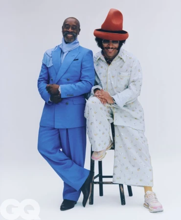Don Cheadle in a blue suit and Boots Riley in a patterned outfit with a red hat posing against a white background for GQ Magazine.