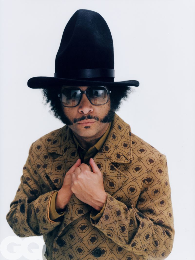 Boots Riley wearing a black hat, tinted sunglasses, and a patterned tan jacket against a plain white background for GQ Magazine U.S.