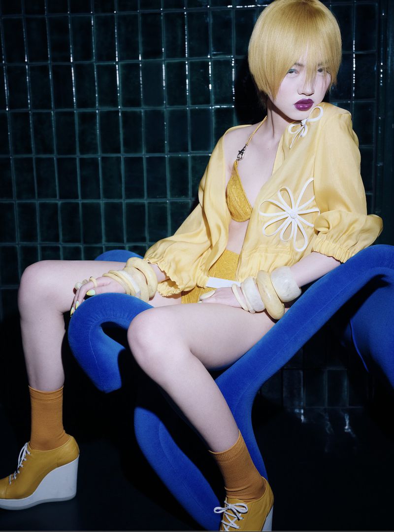 Model with short yellow hair wearing yellow outfit sitting on a blue chair against a dark tiled background