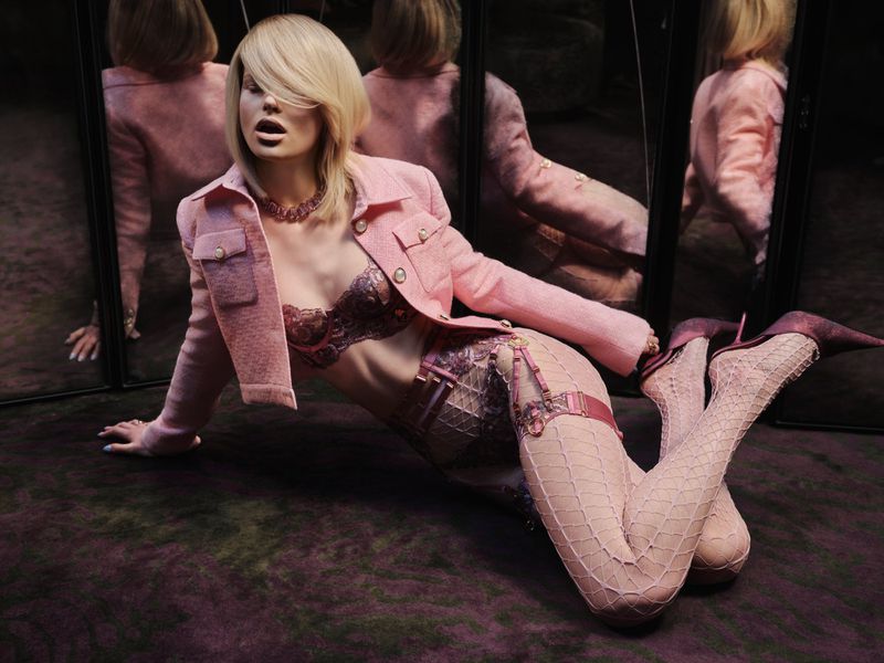 A blond woman wearing a pink tweed jacket and patterned lingerie reclines against mirrors reflecting her figure.