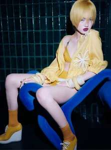Model with short yellow hair wearing yellow outfit sitting on a blue chair against a dark tiled background