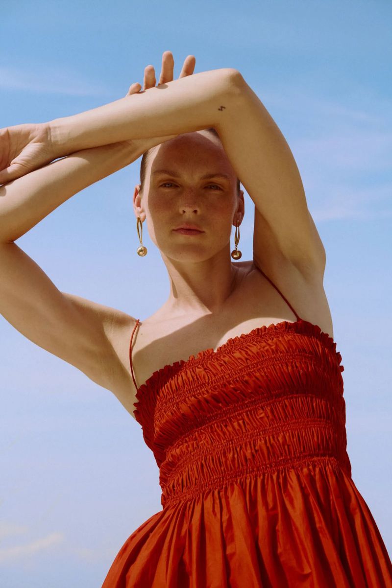 A woman in a red dress poses against a bright sky, arms raised above her head. She wears long gold earrings.