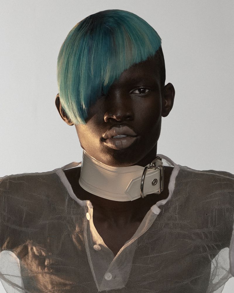 Model with striking blue side-swept hair wearing a white buckled neck collar and sheer textured top against a neutral background.