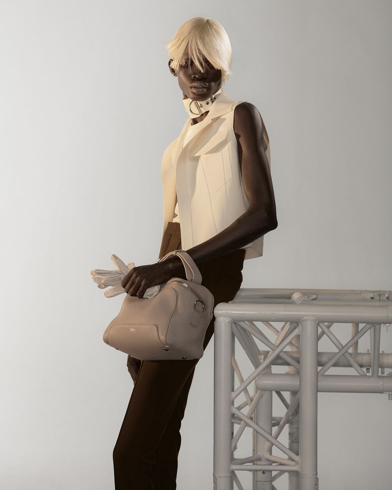 Model with platinum blonde hair wearing a white vest and dark trousers holding a handbag against a plain background