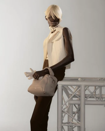 Model with platinum blonde hair wearing a white vest and dark trousers holding a handbag against a plain background