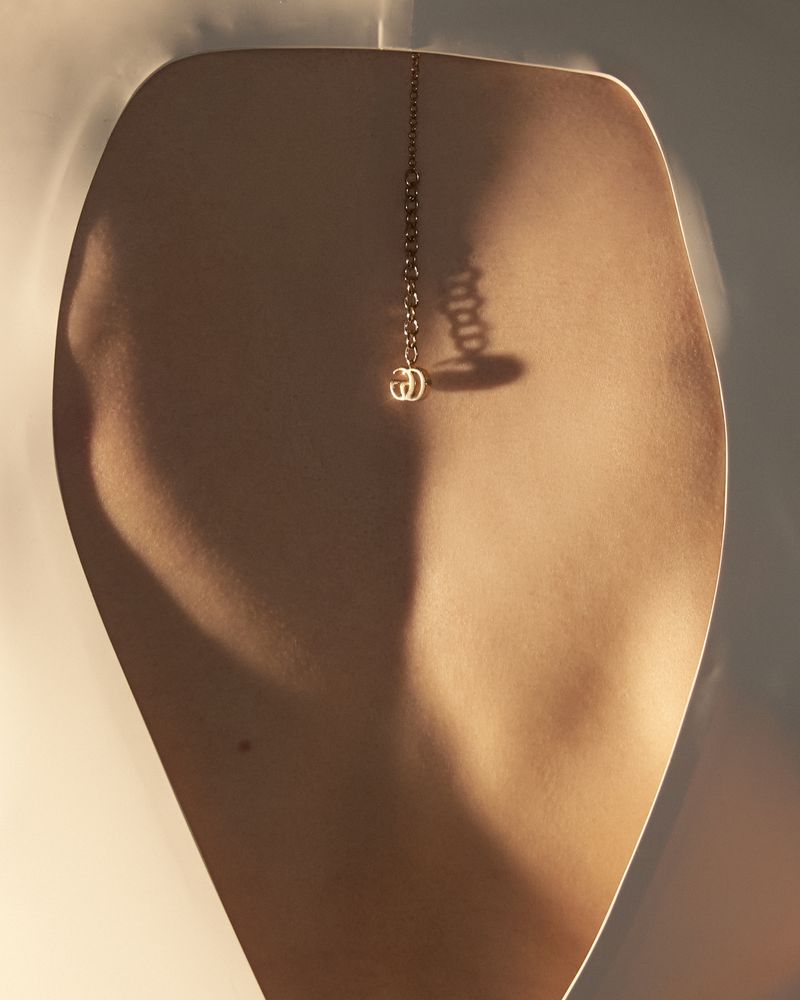 Close-up jewellery product shot of a thin gold necklace with an interlocking logo pendant resting on a sculptural form