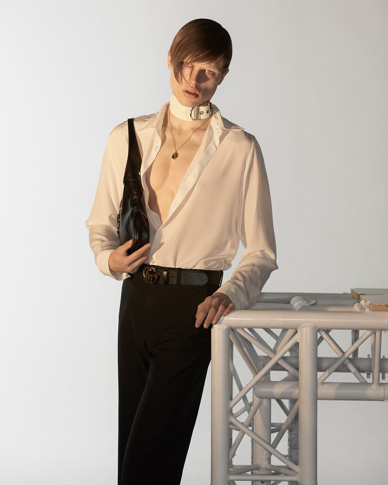 Model wearing a white shirt and black trousers holding a black handbag against a seamless white background.