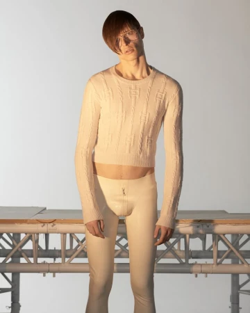 Model wearing cream knit sweater and tight trousers posing against an industrial metal scaffolding background