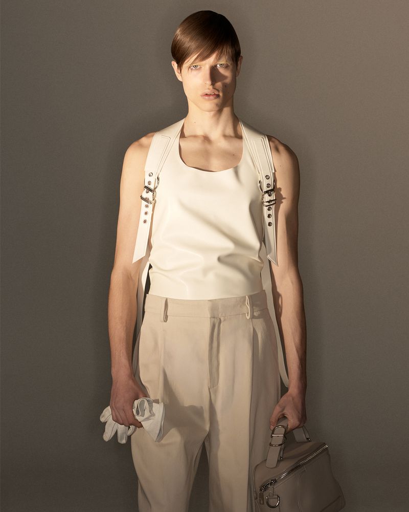 Model wearing a white leather tank top and beige trousers holding gloves and a handbag against a grey backdrop