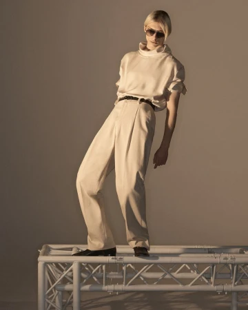 Model wearing tinted sunglasses, cream silk top, and trousers standing on a white structural frame.