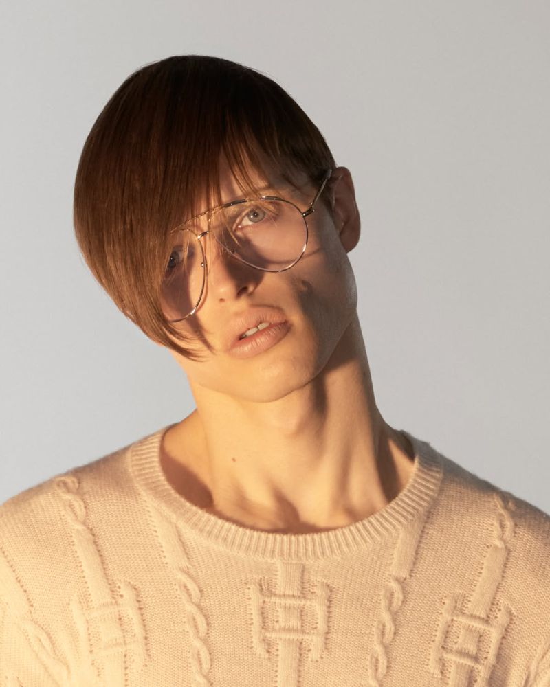 Portrait of a model wearing clear glasses and a patterned cream crewneck sweater against a plain white background.