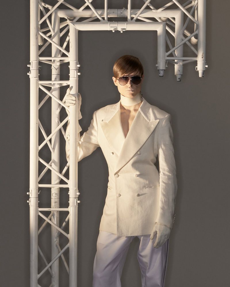 Adult posing with an industrial metal structure wearing a double-breasted white suit, sunglasses, and gloves in a studio setting.