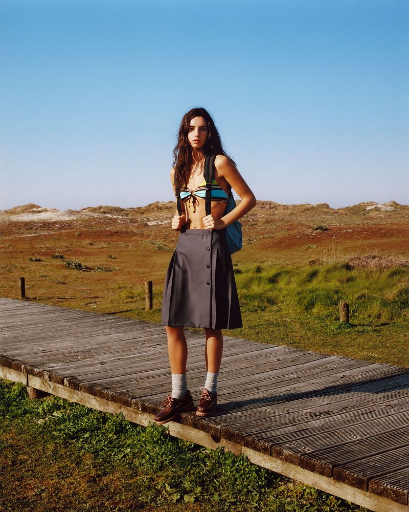 A young woman standing on a wooden boardwalk wearing a pleated grey skirt, bikini top, and backpack with a coastal dune background.