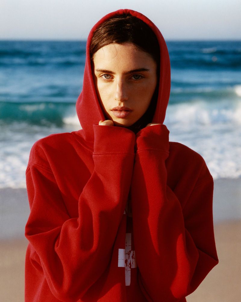 A woman wearing a red hooded sweatshirt standing on a sandy beach by the ocean.