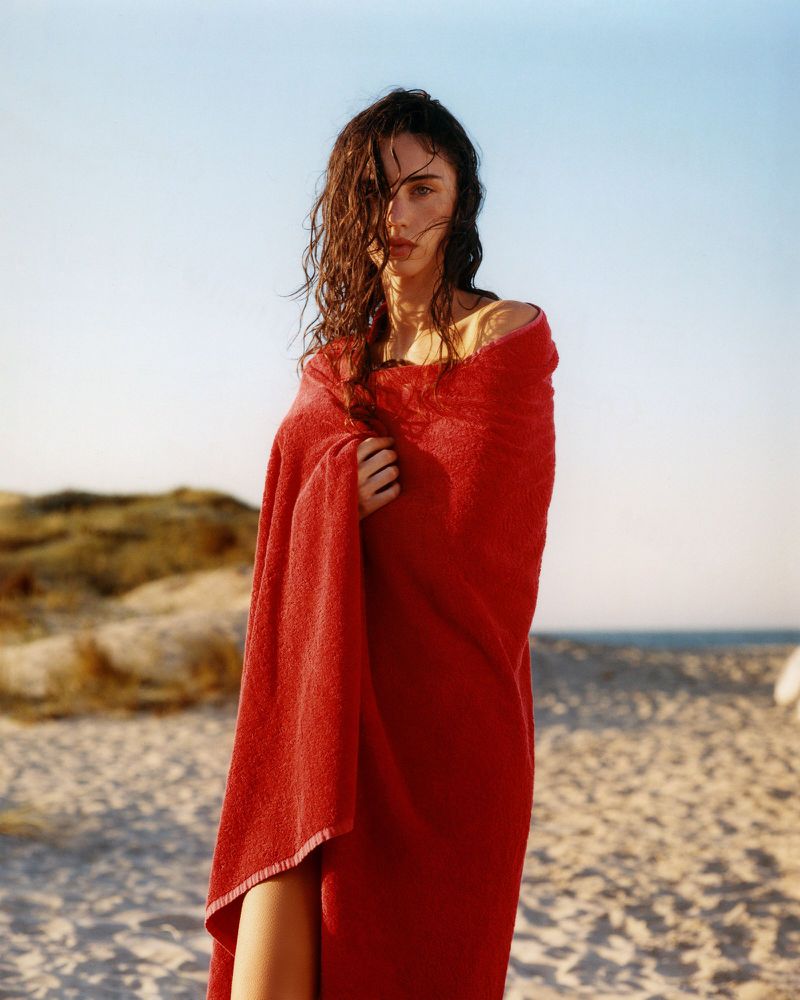 Woman wrapped in a bright red towel standing on a sandy beach under a clear sky.