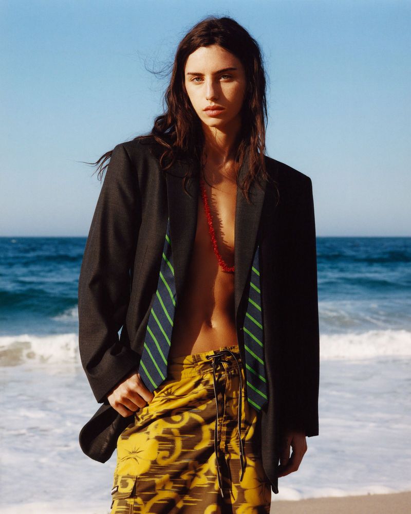Model standing on a sandy beach wearing an oversized black blazer and yellow patterned shorts against an ocean backdrop.