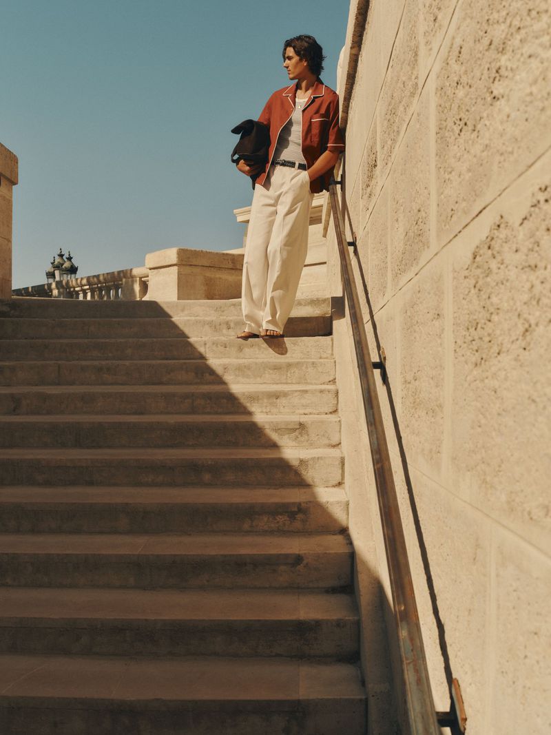 Man standing on stone steps leaning against a railing wearing a terracotta shirt and white trousers under clear sky