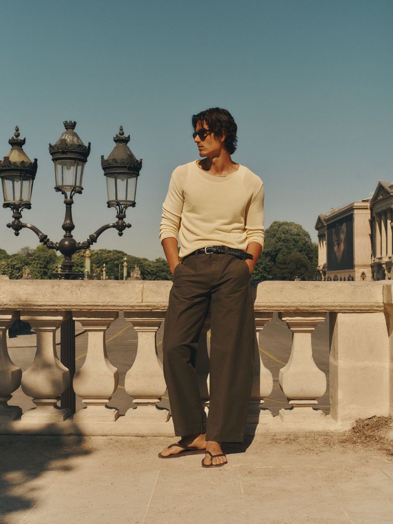 Man wearing cream knit sweater and dark trousers leaning against a stone balustrade outdoors.