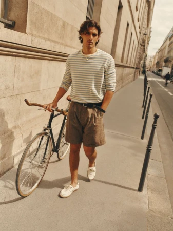 Man wearing a striped long-sleeve shirt and brown shorts standing with a bicycle on a sunlit city street