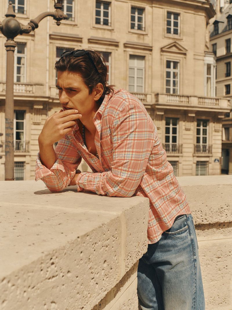 Man wearing a pink plaid button-down shirt and blue denim jeans leaning on a stone railing with a classic building in the background.