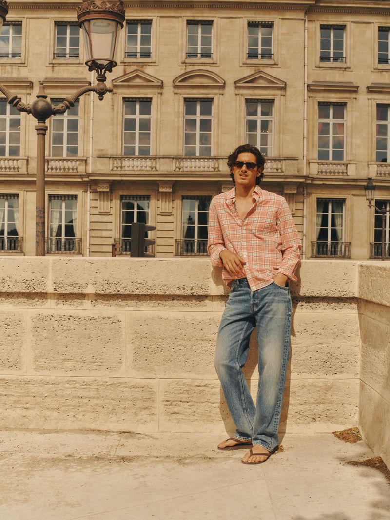 Man wearing a pink plaid shirt and blue jeans leaning against a stone wall in front of a classical building