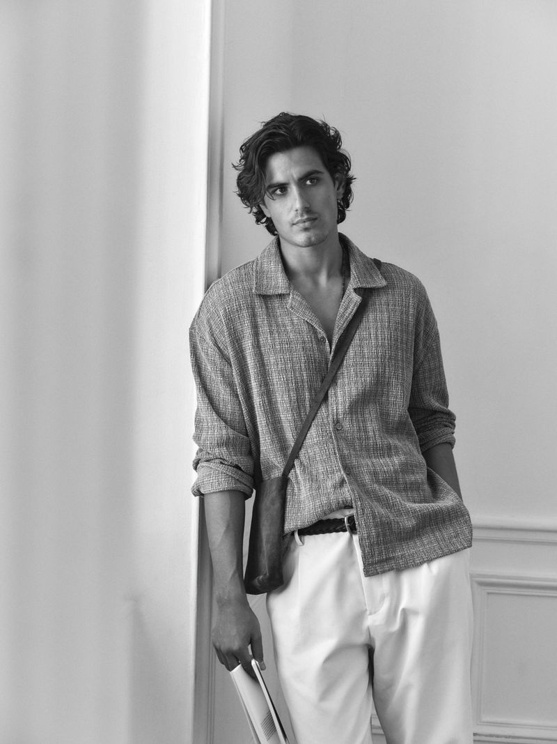 Black and white shot of a male model wearing an oversized collared shirt, white pants, and a shoulder bag, standing indoors against a white wall.