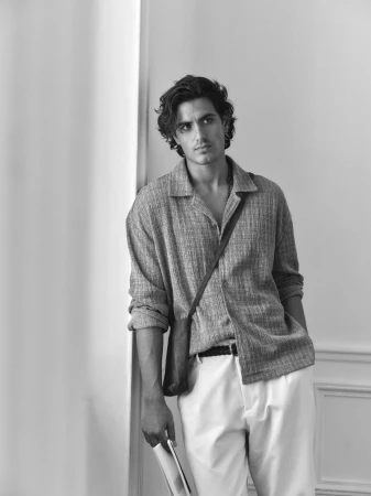 Black and white shot of a male model wearing an oversized collared shirt, white pants, and a shoulder bag, standing indoors against a white wall.