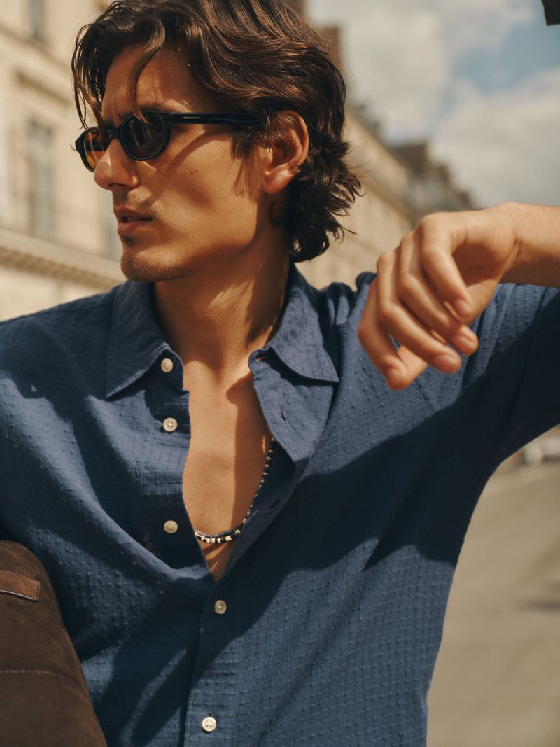 Man wearing sunglasses and a blue textured shirt leaning against an outdoor structure in a street setting.