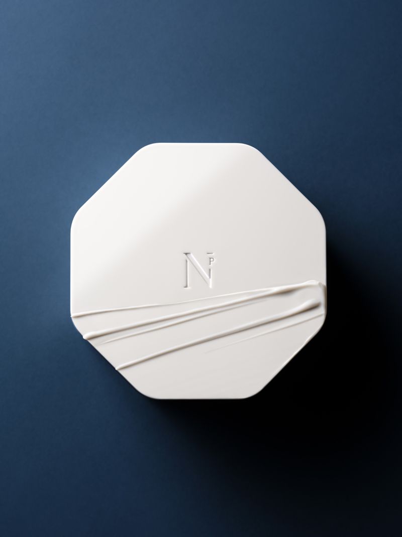 Close-up of an octagonal-shaped white cosmetic compact case with a cream swatch on a solid dark blue background, eye-level view.