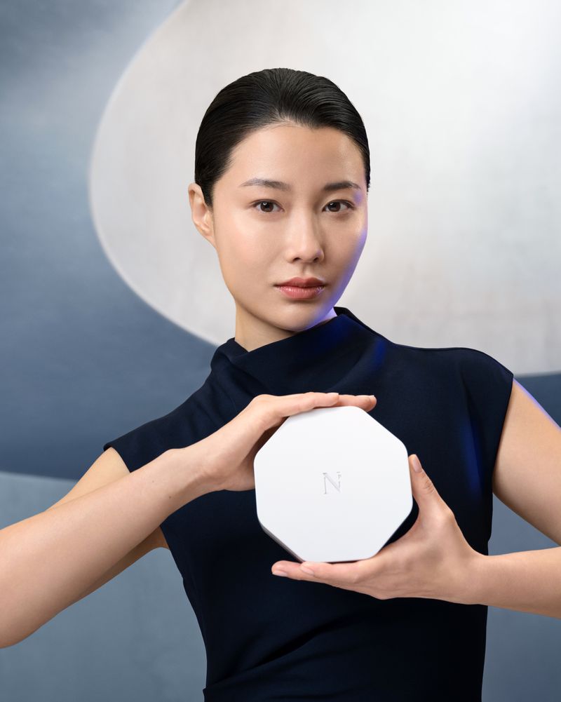 Asian woman holding white octagonal box in navy dress against blue and white patterned backdrop in a beauty and fashion shot.