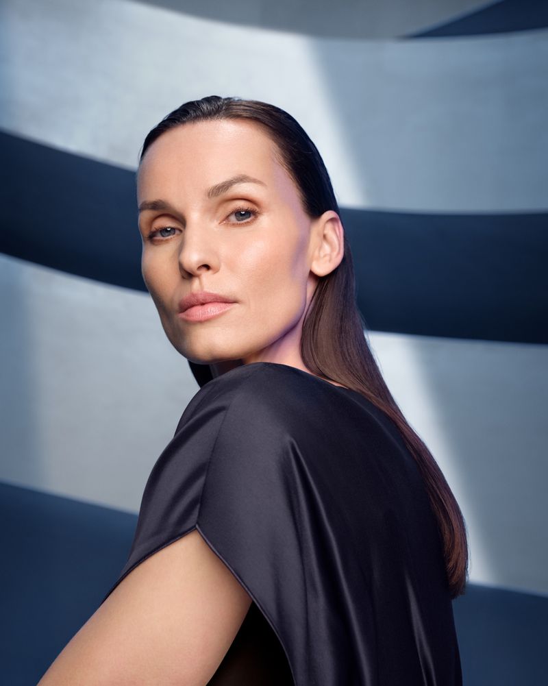 Portrait of an adult woman with brown hair looking over her shoulder in a black dress against a graphic blue and white background.