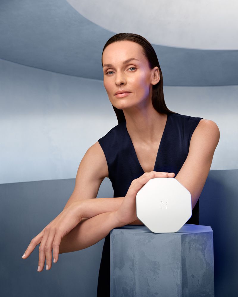 Adult female with dark hair poses indoors while holding a white cosmetic compact on top of a cubic blue pillar with a geometric background.