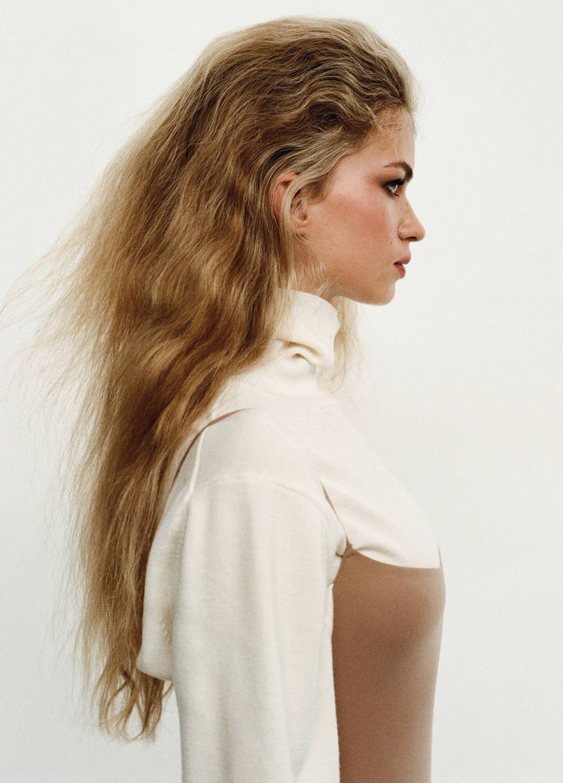 Profile portrait of a woman with long wavy blonde hair wearing a white turtleneck top against a plain light background