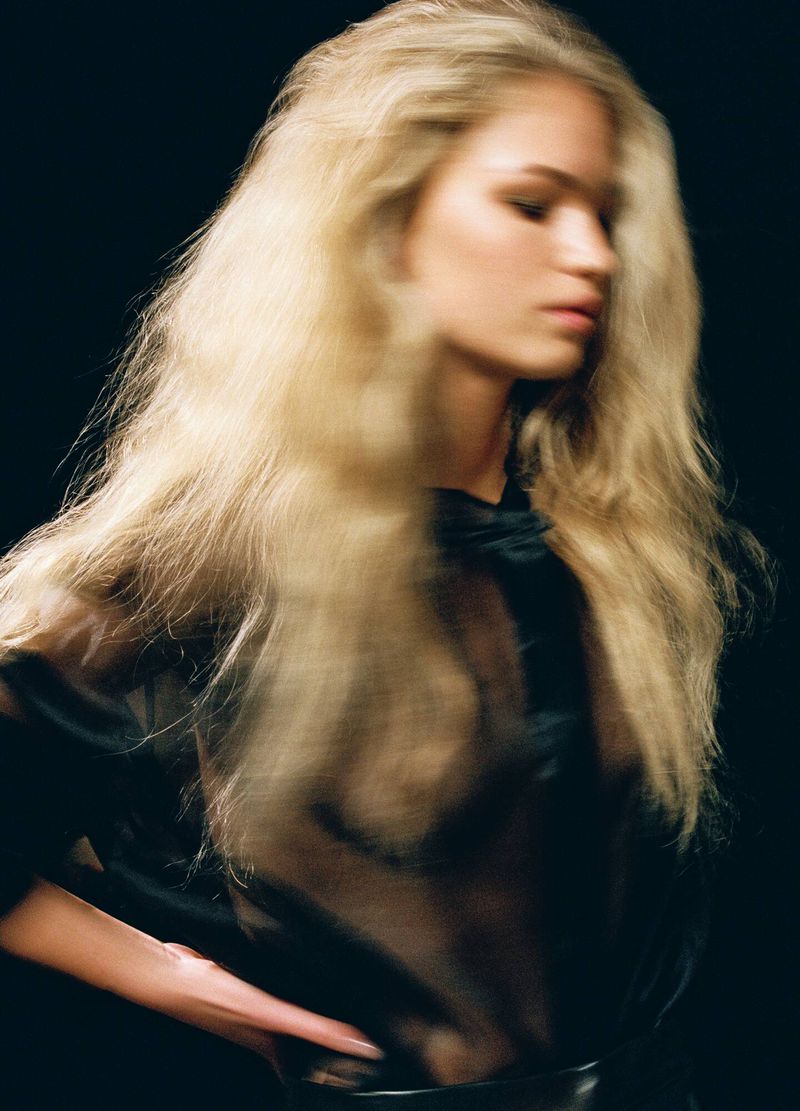Portrait of a blonde model with long tousled hair wearing a sheer black top against a dark background.