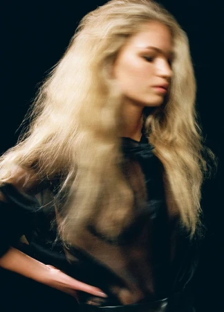 Portrait of a blonde model with long tousled hair wearing a sheer black top against a dark background.
