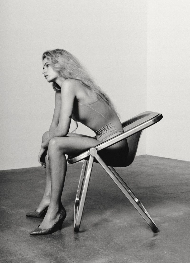 A young fair-skinned woman with long blond hair, wearing a bathing suit and high heels, is sitting on a chrome folding chair indoors.
