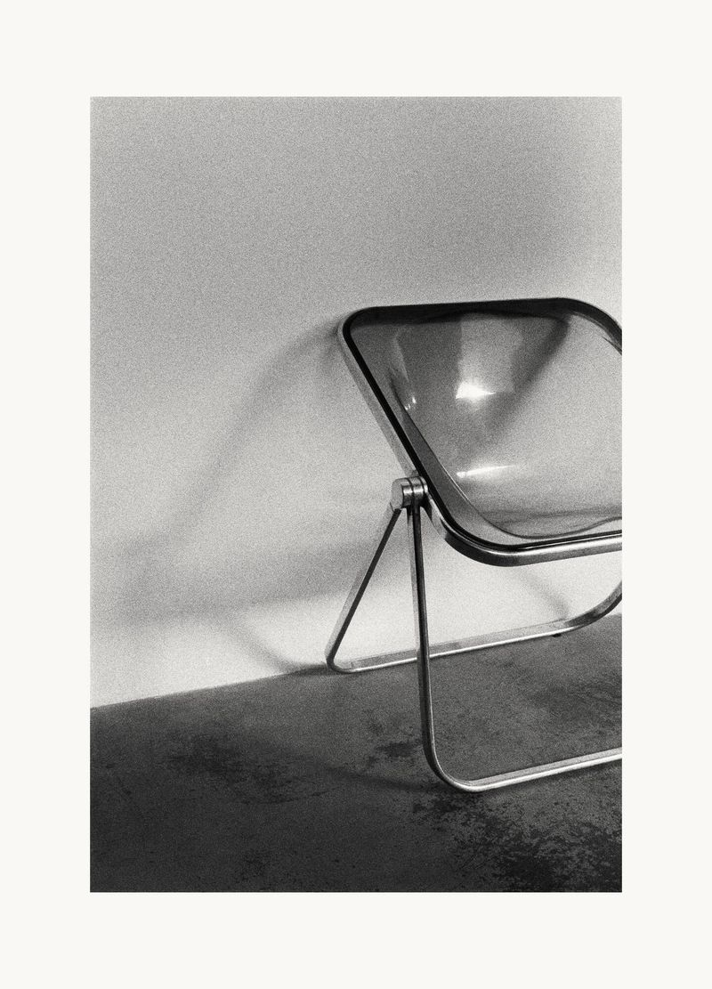 Transparent armchair with a metal frame positioned against a plain wall in a monochrome photograph.