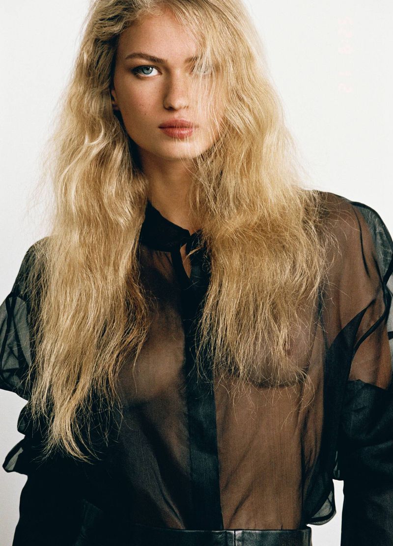 Portrait of a blonde woman wearing a sheer black blouse against a plain light background