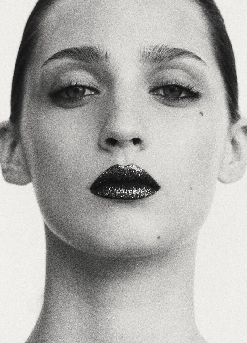 Close-up portrait of a woman with dark sparkly lipstick and dark eyeliner against a seamless white background.