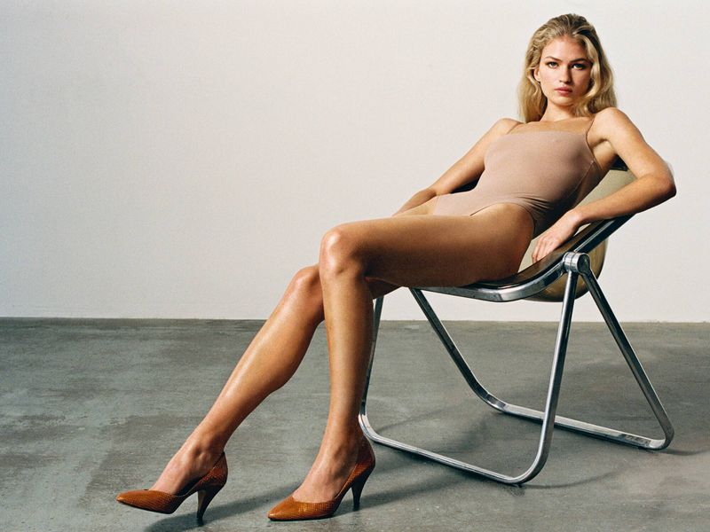 Model wearing a beige bodysuit and brown heels reclining in a metal chair against a neutral background.