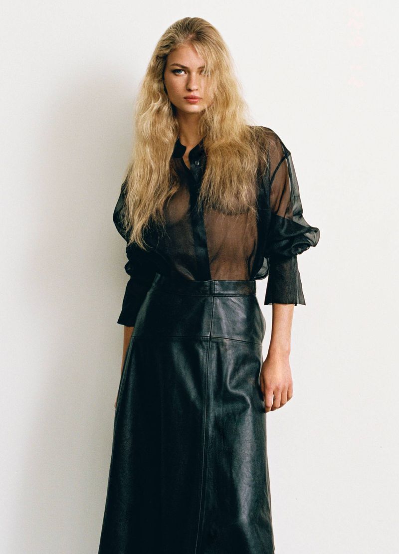 Model with long blonde hair wearing a black sheer blouse and a black leather skirt against a white studio background.