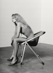 Monochrome fashion portrait of a model with long hair seated sideways on a modern metal tubular chair.