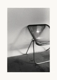Transparent armchair with a metal frame positioned against a plain wall in a monochrome photograph.