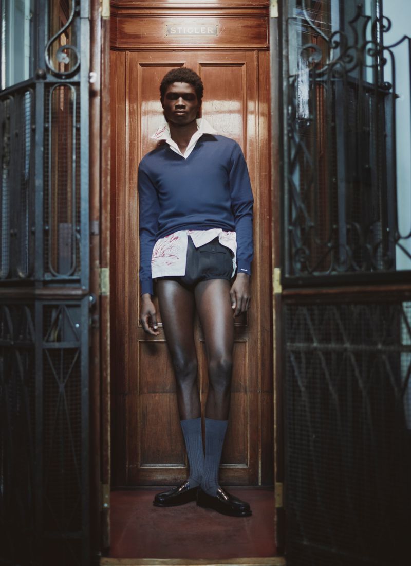 Full-length portrait of a male model posing in a navy sweater and shorts standing in a wooden doorway with wrought-iron grates.