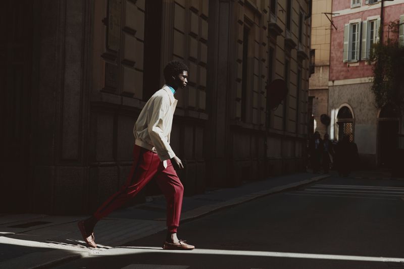 A man walking across a sunlit European street wearing a light jacket and red trousers, photographed for ICON Magazine Mena.
