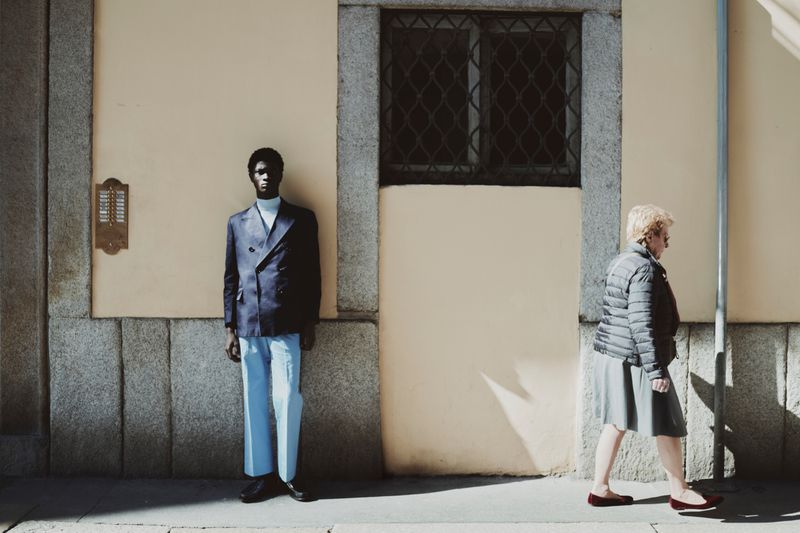 A man in a navy double-breasted blazer and light blue trousers stands against a wall as a woman walks past in profile.